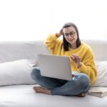 stylish-brunette-girl-yellow-sweater-sits-home-sofa-bright-room-works-laptop-remotely_169016-10242